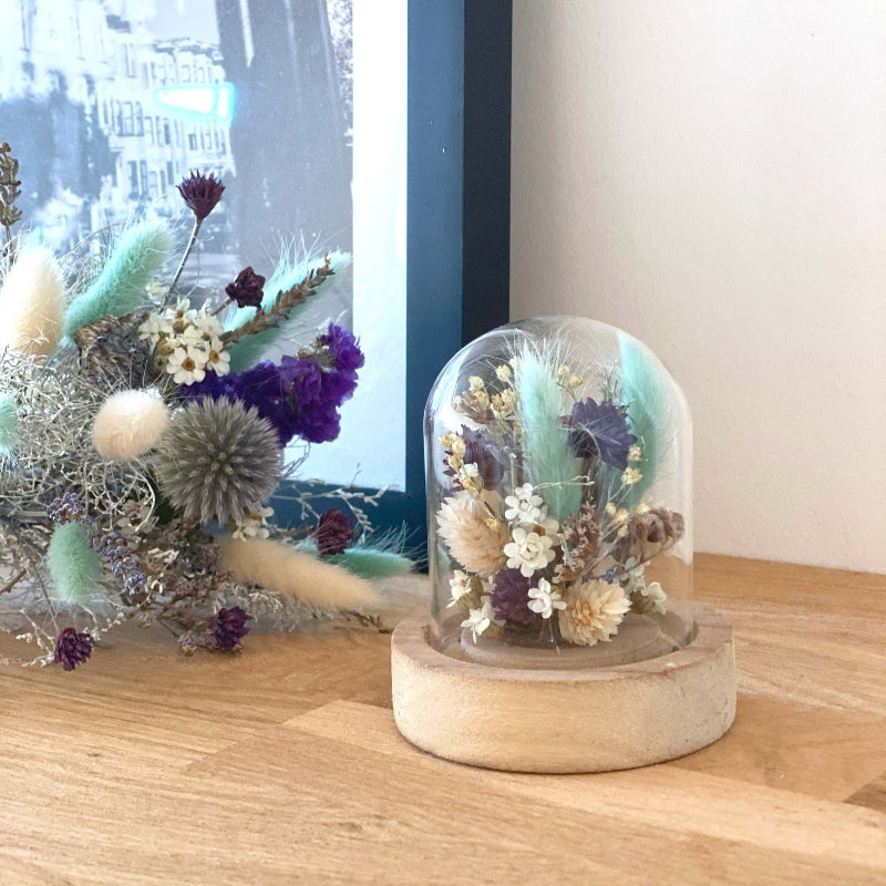 Composition de fleurs sechees dans les couleurs blanc bleu et violet delicatement arrangees sous un globe en verre