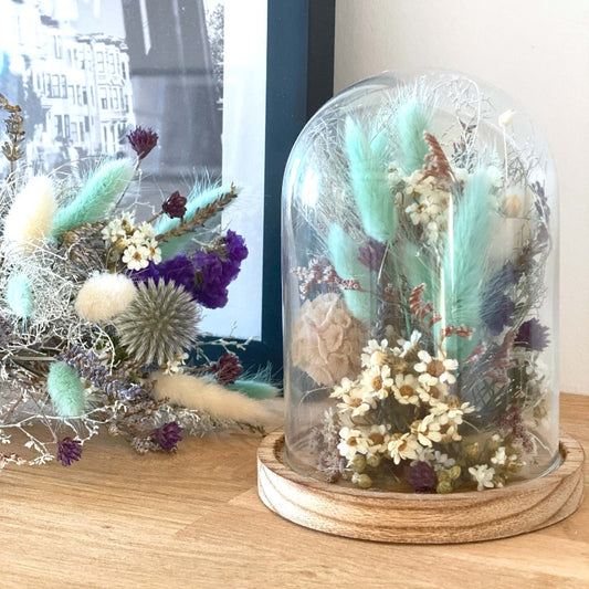 Composition de fleurs sechees dans les couleurs blanc bleu et violet delicatement arrangees sous un globe en verre