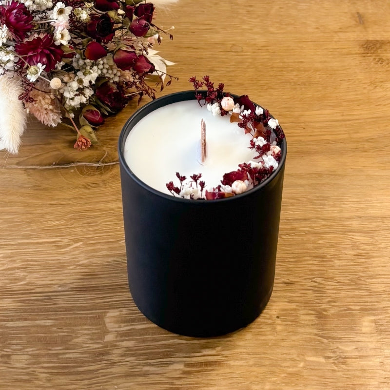 bougie composée de cire naturelle, décorée de petites fleurs séchées dans les tons rouge foncé et blanc, avec mèche en bois, coulée dans un contenant en verre noir mat